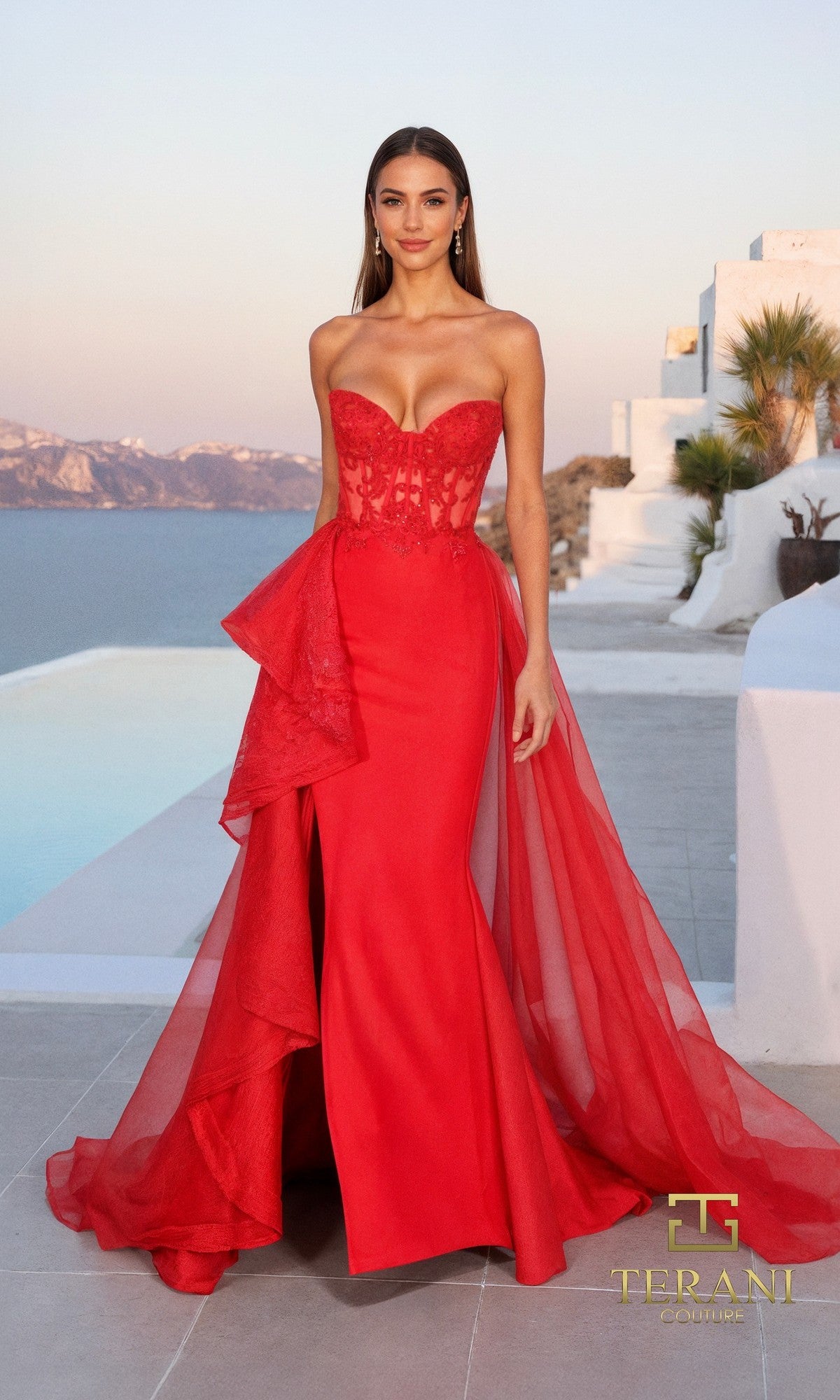 A woman stands by a pool in the red Terani 261P6261 long formal dress. The strapless Mikado gown features lace, sheer details, and a flowing tulle overskirt, set against coastal scenery and white buildings for chic elegance.